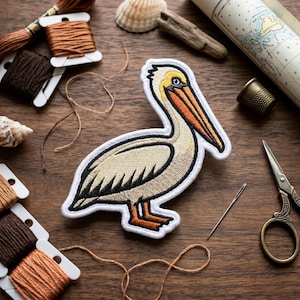 May include: An embroidered patch featuring a pelican with cream-colored feathers, a long orange beak, and orange feet. The patch has a thick white border and is surrounded by various sewing supplies including spools of brown thread and scissors.