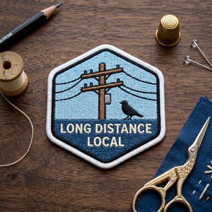 May include: A hexagonal embroidered patch featuring a light blue sky background, a brown utility pole with black power lines, and a black bird perched on the ground. The dark blue lower section contains the white text LONG DISTANCE LOCAL.