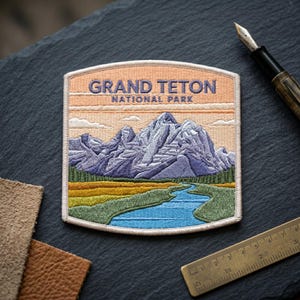 May include: A rectangular embroidered patch featuring a landscape of the Grand Teton National Park with purple mountains, a blue river, and green fields under a peach sky. The text reads GRAND TETON NATIONAL PARK in dark blue lettering.