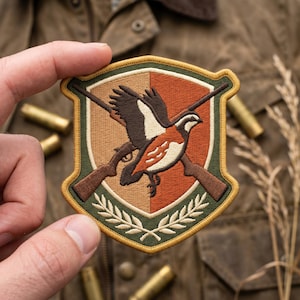 May include: A shield-shaped embroidered patch featuring a flying game bird in flight over two crossed brown shotguns. The background is split between tan and orange, framed by a dark green border with a gold edge and a laurel leaf design.