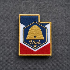 May include: A rectangular embroidered patch shaped like the state of Utah. The design features a blue, white, and red background with a central navy blue hexagon containing a gold beehive icon, the word Utah in script, and a small gold star.