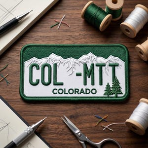 May include: A rectangular embroidered patch featuring a white background with a green border. The design displays white snow-capped mountains above the dark green text COL-MTT and COLORADO, with two small green pine trees on the right side.