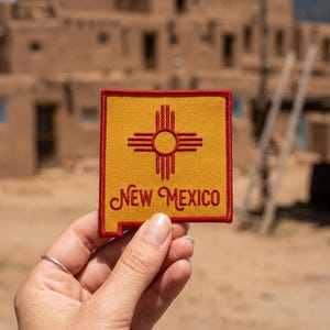 May include: A square-shaped embroidered patch featuring a yellow background with a red border and a red Zia sun symbol centered above the red embroidered text that reads New Mexico in a cursive script font.