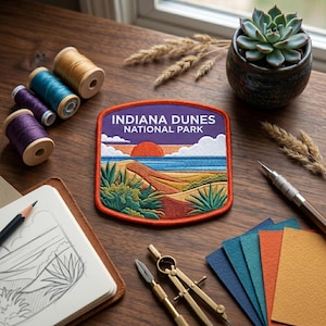 May include: An embroidered patch featuring a landscape of Indiana Dunes National Park with a red sun, blue water, and sand dunes. The text reads INDIANA DUNES NATIONAL PARK. The patch has a thick orange border and is surrounded by sewing supplies.