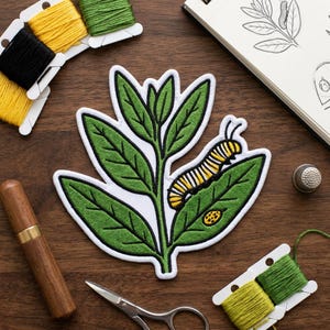 May include: Embroidered patch featuring a green leaf with a caterpillar and ladybug. The patch is white-bordered and surrounded by spools of yellow, black, and green thread, a needle case, scissors, a thimble, and a sketchbook on a wooden surface.