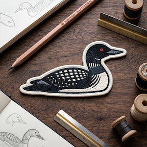 May include: An embroidered patch of a common loon featuring black and white plumage with a distinct red eye. The patch is positioned on a dark wood surface alongside a wooden pencil, two metal rulers, and several wooden spools of thread.