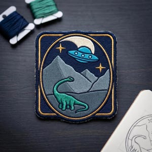 May include: Embroidered patch featuring a teal dinosaur under a blue UFO, set against a mountain range and starry sky. The patch has a navy blue border with gold accents. Threads in teal and navy are visible.