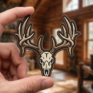 May include: An embroidered patch featuring a deer skull with large, branching antlers. The skull is cream-colored with black outlines, while the antlers are shaded in light tan and brown tones. The entire design is bordered by a dark brown thread.