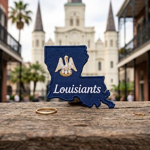 May include: A dark blue embroidered patch shaped like the state of Louisiana featuring a white and gold pelican emblem above the white cursive text Louisiants, accompanied by a thin gold metal ring resting on a weathered wooden surface.
