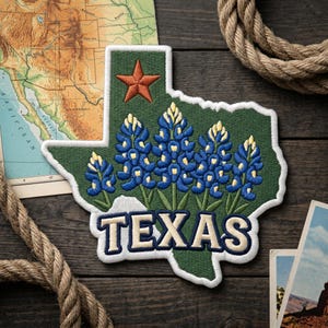 May include: Embroidered patch in the shape of Texas, featuring bluebonnets and a star. The word "TEXAS" is embroidered in white with a navy blue outline. The patch is on a dark wooden surface with a map and rope.
