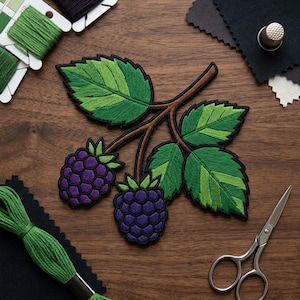 May include: Embroidered patch featuring a blackberry branch with two purple berries and green leaves. The leaves have a two-tone green design. The patch is on a wooden surface with spools of green thread, scissors, and fabric swatches.