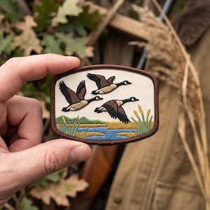 May include: A rectangular embroidered patch with a dark brown border featuring three geese in flight over a landscape of blue water, yellow marshland, and green reeds against a cream-colored background. The design is detailed with thread.