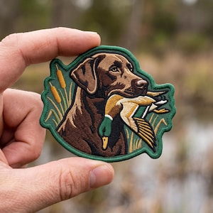 May include: An embroidered patch featuring a brown Labrador retriever holding a mallard duck in its mouth. The design includes yellow cattails and green reeds against a dark green border, showcasing detailed stitching of the dog and bird.