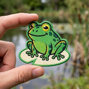 May include: An embroidered patch featuring a bright green frog with dark green spots and yellow accents, sitting on a light beige lily pad. The frog has large yellow eyes with black pupils and a wide, smiling mouth, all outlined in thick black thread.