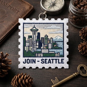 May include: Embroidered patch featuring the Seattle skyline, including the Space Needle, with the text "JOIN - SEATTLE." The patch is white with a blue border and a postage stamp design. Other items in the image include a compass, passport, and coffee beans.