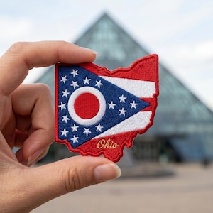 May include: An embroidered patch shaped like the state of Ohio featuring the state flag design. The patch displays a blue triangle with a white-bordered red circle and seventeen white stars, alongside red and white stripes, with the word Ohio in gold.