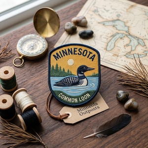 May include: A Minnesota patch featuring a Common Loon swimming on a lake, with a yellow sky and green trees. The patch is on a wooden surface with a compass, map, rocks, and spools of thread.