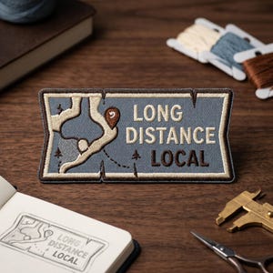 May include: A rectangular embroidered patch with a blue-grey background, featuring a cream-colored winding path, a brown location pin, and the text "LONG DISTANCE LOCAL" in cream and brown thread, all bordered by a dark brown stitched edge.