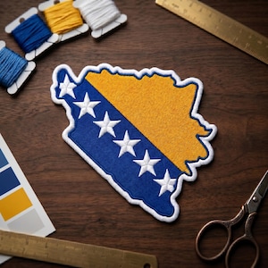 May include: Embroidered patch in the shape of Bosnia and Herzegovina, featuring the country's flag design. The patch has a blue and yellow color scheme with white stars. Sewing supplies, including thread spools and scissors, are arranged around the patch.