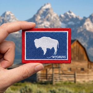 May include: A rectangular embroidered patch featuring the Wyoming state flag design. The patch has a blue field with a white silhouette of an American bison in the center, a white border, and a red outer border. The word WYOMING is stitched in white.