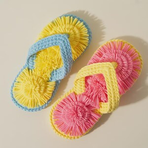 以下が含まれることがあります： 手作りのかぎ針編みビーチサンダルのペア。片方は黄色と青、もう片方はピンクと黄色です。ビーチサンダルは、フットベッドとストラップにテクスチャード加工されたフリンジデザインが施されています。