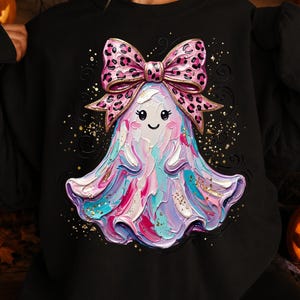May include: A black crewneck sweatshirt featuring a central graphic of a ghost with a textured, impasto-style paint effect in shades of pink, blue, and white. The ghost wears a large pink and black leopard print bow and is surrounded by gold glitter.