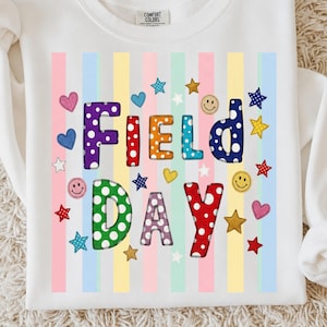 Può includere: Felpa bianca con la scritta "FIELD DAY" in lettere colorate e decorate. Il design include cuori, stelle e faccine sorridenti, su uno sfondo di strisce verticali pastello. È visibile un'etichetta Comfort Colors.