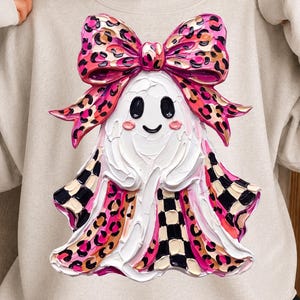 May include: A light beige crewneck sweatshirt featuring a central graphic of a white ghost with a thick, textured paint effect. The ghost wears a large pink leopard print bow and has a skirt pattern of pink leopard print and black and white checkers.