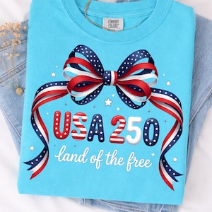 Puede incluir: Una sudadera azul claro con un diseño patriótico. El diseño presenta un lazo hecho de cintas rojas, blancas y azules, el texto "USA 250" y la frase "land of the free". La sudadera está doblada sobre un par de vaqueros azules.