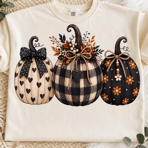 May include: A cream-colored t-shirt featuring a graphic of three pumpkins. The left pumpkin is cream with black hearts and a polka-dot bow. The center pumpkin has a black and white buffalo check pattern with autumn foliage. The right pumpkin is black with orange flowers.