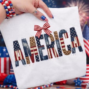 May include: White t-shirt with the word "AMERICA" in a patchwork design. The letters feature denim, leopard print, and the American flag. A red and white striped bow is at the top. Patriotic apparel.