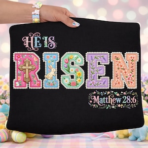 Könnte beinhalten: Schwarzes Kissen mit dem Schriftzug "He Is Risen" in bunten, floral verzierten Buchstaben. Der Buchstabe "R" zeigt ein Kreuz, und der Buchstabe "I" einen Hasen. Der Text "Matthew 28:6" steht unter dem Haupttext.