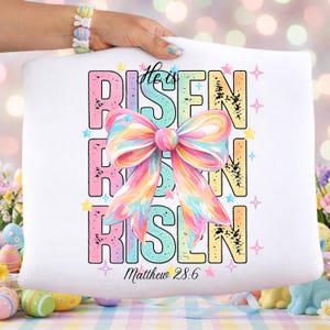 May include: White pillow with the words "He is Risen" in pastel colors, with a large, colorful bow in the center. The text "Matthew 28:6" is at the bottom, with star accents. The pillow is held by a person.