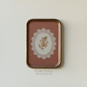 May include: A framed wall art piece featuring a floral design. The artwork has a brown background with an oval-shaped lace doily in the center, showcasing a delicate pink floral arrangement. The frame is a warm, metallic gold color. The text "FLORA VETUS HERITAGE WALL ART" is at the bottom.