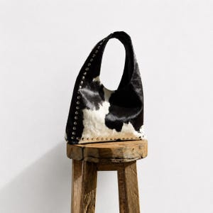 May include: A hobo-style handbag crafted from black and white cowhide leather featuring a distinctive spotted pattern. The bag is accented with a vertical row of silver-toned metal studs along the side and a matching row of studs along the base.
