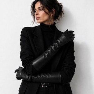 May include: A pair of long black leather gloves reaching past the elbows, worn with a black turtleneck sweater and a black blazer. The gloves have a smooth, matte finish and a fitted silhouette that extends up the forearm, providing full coverage.