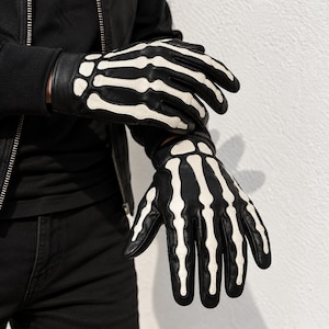 May include: A pair of black leather gloves featuring a white skeleton hand bone design printed on the back of the hands and fingers. The gloves have a smooth finish and cover the fingers and palms, extending to the wrists with a fitted cuff.