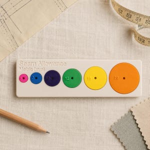 Puede incluir: Una guía de margen de costura rectangular de color beige con el texto "Seam Allowance Metric (mm)" y seis recortes circulares de colores etiquetados con 5, 7, 10, 12, 15 y 20. La herramienta incluye círculos rosa, azul, morado, verde, amarillo y naranja.