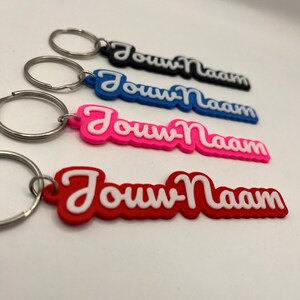 Op de afbeelding: Vier sleutelhangers met de tekst "Jouw Naam" in een cursief lettertype. De sleutelhangers zijn zwart, blauw, roze en rood, elk met een witte rand. De sleutelhangers hebben zilveren sleutelringen.