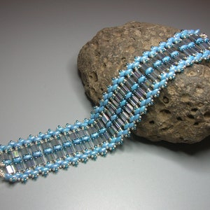 May include: A blue and silver beaded bracelet with a toggle clasp. The bracelet is made with small, round beads and larger, rectangular beads.