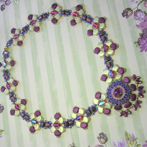 Op de afbeelding: Een paarse en witte kralenketting met een bloemenpatroon. De ketting heeft een grote bloemhanger in het midden.