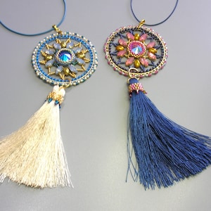 Puede incluir: Dos collares de cuentas con colgantes redondos, coloridos, con forma de mandala y borlas. Una borla es blanca y la otra es azul.