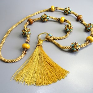 Può includere: Una collana di perline dorate con un fiocco e perline turchesi e marroni. La collana è fatta di piccole perline dorate e ha un fiocco alla fine.