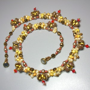 Pode incluir: Um colar de várias voltas com contas marrons, douradas e brancas. O colar tem detalhes em cristal vermelho e um fecho.