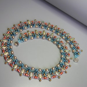 Puede incluir: Un collar de cuentas de m&uacute;ltiples hilos con un patr&oacute;n geom&eacute;trico en tonos de azul verdoso, marr&oacute;n, rosa y blanco. El collar tiene un cierre magn&eacute;tico.