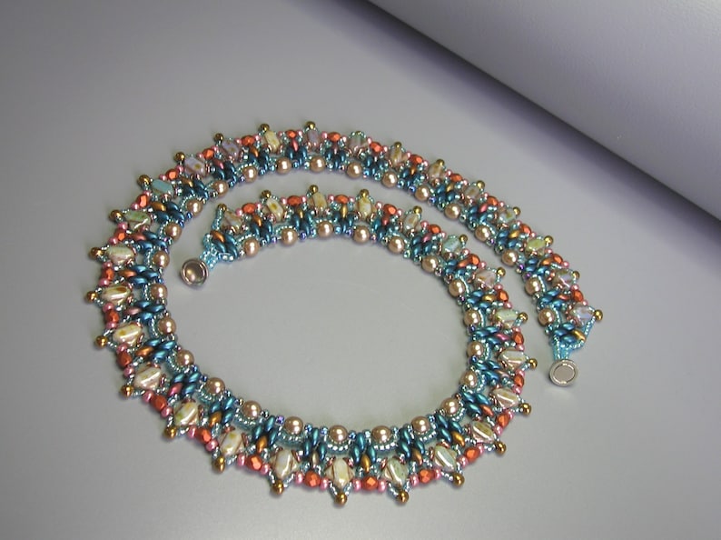 Puede incluir: Un collar de cuentas de m&uacute;ltiples hilos con un patr&oacute;n geom&eacute;trico en tonos de azul verdoso, marr&oacute;n, rosa y blanco. El collar tiene un cierre magn&eacute;tico.