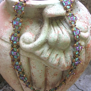 May include: A beaded necklace with purple, turquoise, and gold accents. The necklace is draped over a textured, green and brown ceramic vase.