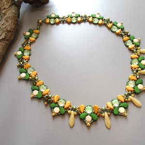 Könnte beinhalten: Eine handgefertigte Halskette mit floralem Design, bestehend aus grünen, gelben und cremefarbenen Perlen. Die Halskette zeigt ein detailliertes Muster mit klaren Kristallakzenten und tropfenförmigen Anhängern. Die Halskette wird auf einer grauen Oberfläche präsentiert.