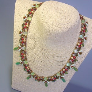 Può includere: Una collana di perline con perline verdi, rosse e dorate. La collana è su un manichino bianco.