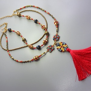 May include: A multi-strand necklace with gold, red, and black beads, and a tassel pendant with a blue and gold floral design. The tassel is red.